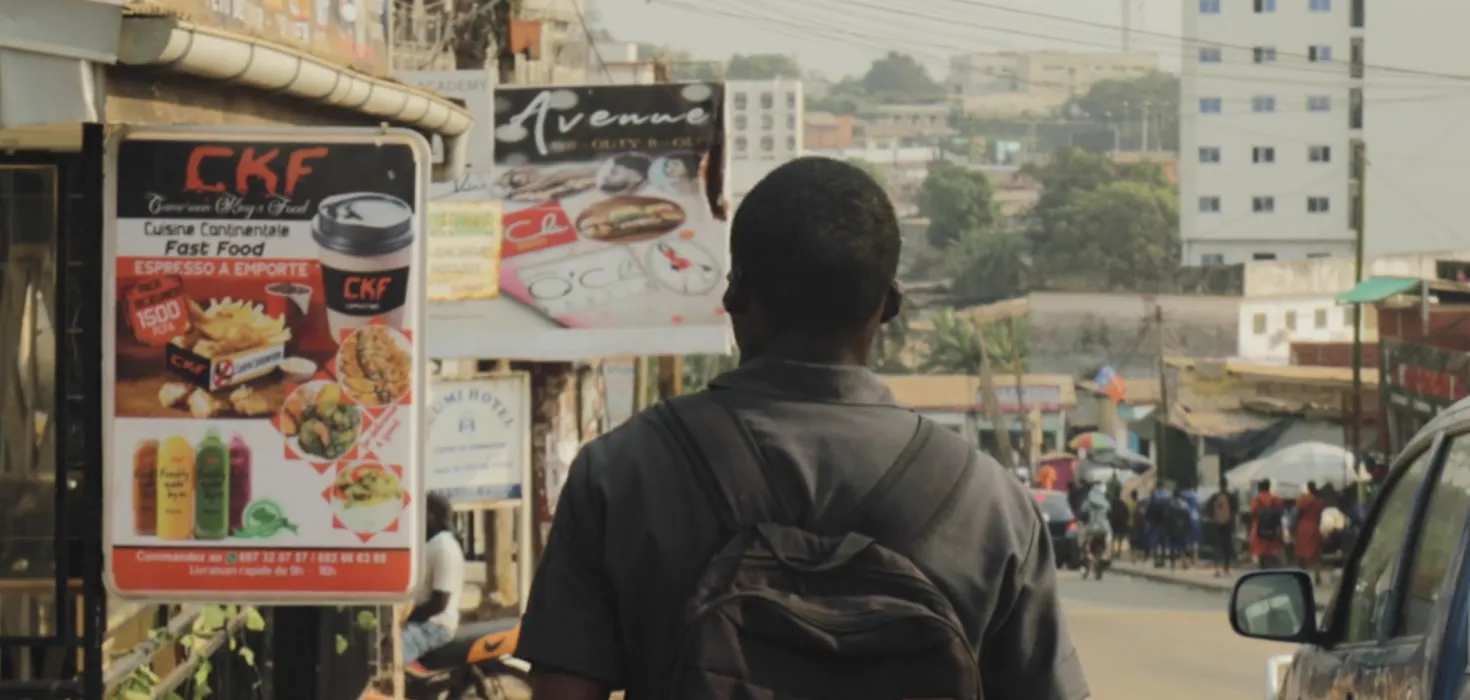 Teenager walks along a street in Yaounde, Cameroon, next to junk food advertisements. Still from 'Africa's diabetes rise' from Turning the Tide series