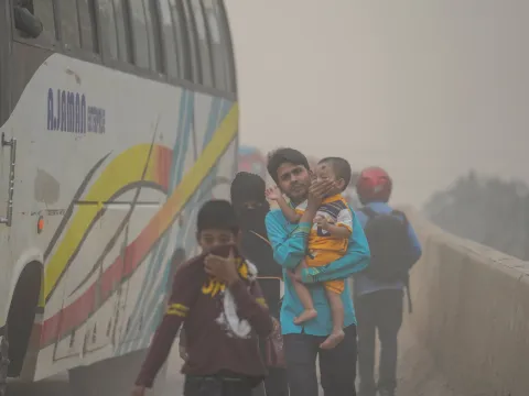A bus on a hazy, polluted road carries several passengers, including a man holding a small child, while others cover their faces from the dust.