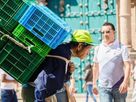 A local peruvian old man works with a heavy load while a tourist walks in the background
