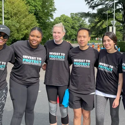 Group of six noncommunicable disease advocates in black t-shirts with 'Invest to Protect' printed on them, standing outdoors during WHO Walk the Talk Event, with trees  in the background.