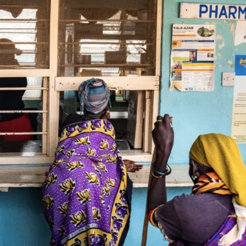 Two women in traditional attire consult at a rural African pharmacy counter.
